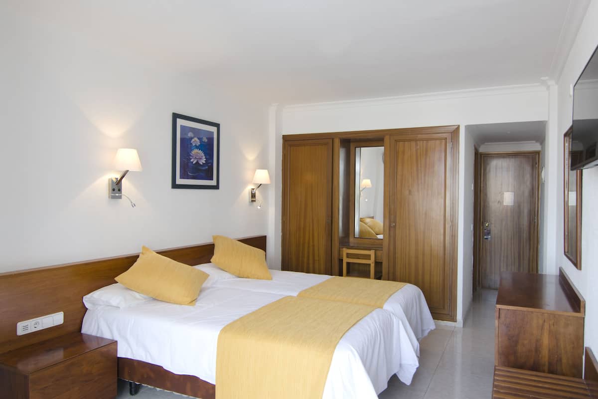 Habitació d'hotel amb un llit gran, amb capçalera de fusta, dues làmpades de sobretaula, una taula de nit, un banc de fusta, un mirall i un armari.