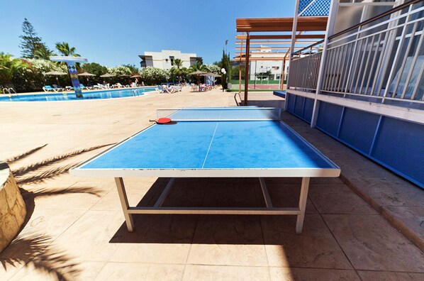 A ping pong table in a sunny outdoor area with a pool and lounge chairs.