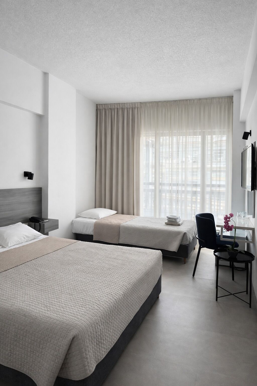 A hotel room with two beds, a desk, a chair, a TV, and a large window with curtains.