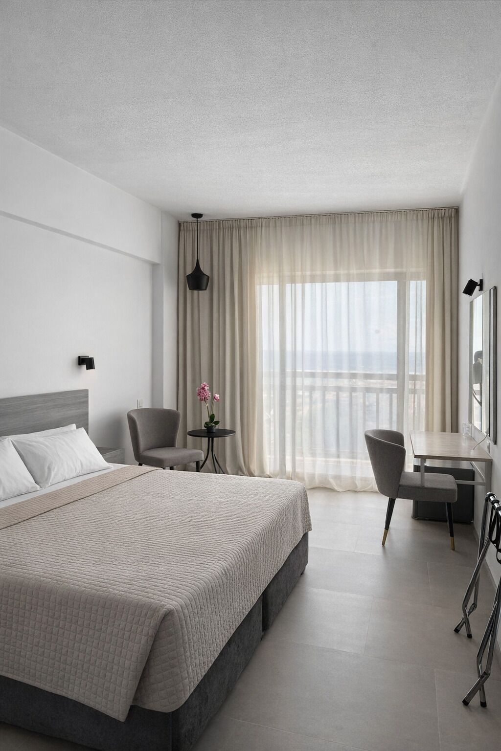A hotel room with a bed, a desk, a chair, a small table with flowers, and a view of the outside.