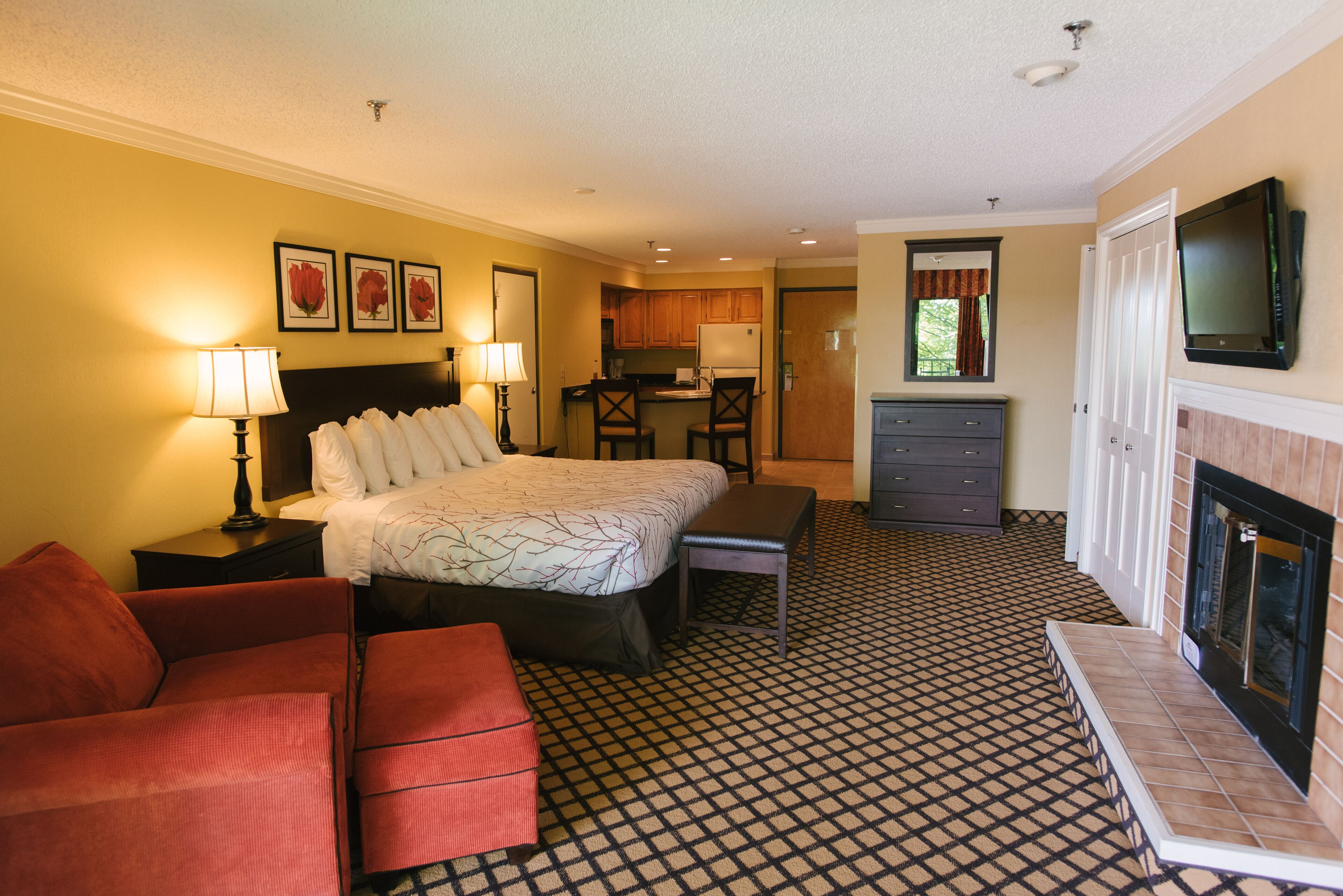 A hotel room with a bed, a desk, a chair, a sofa, a TV, a fireplace, and a kitchen area.