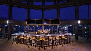 Een barruimte met een centrale bar en meerdere stoelen, grote ramen en een uitzicht op de nachthemel.