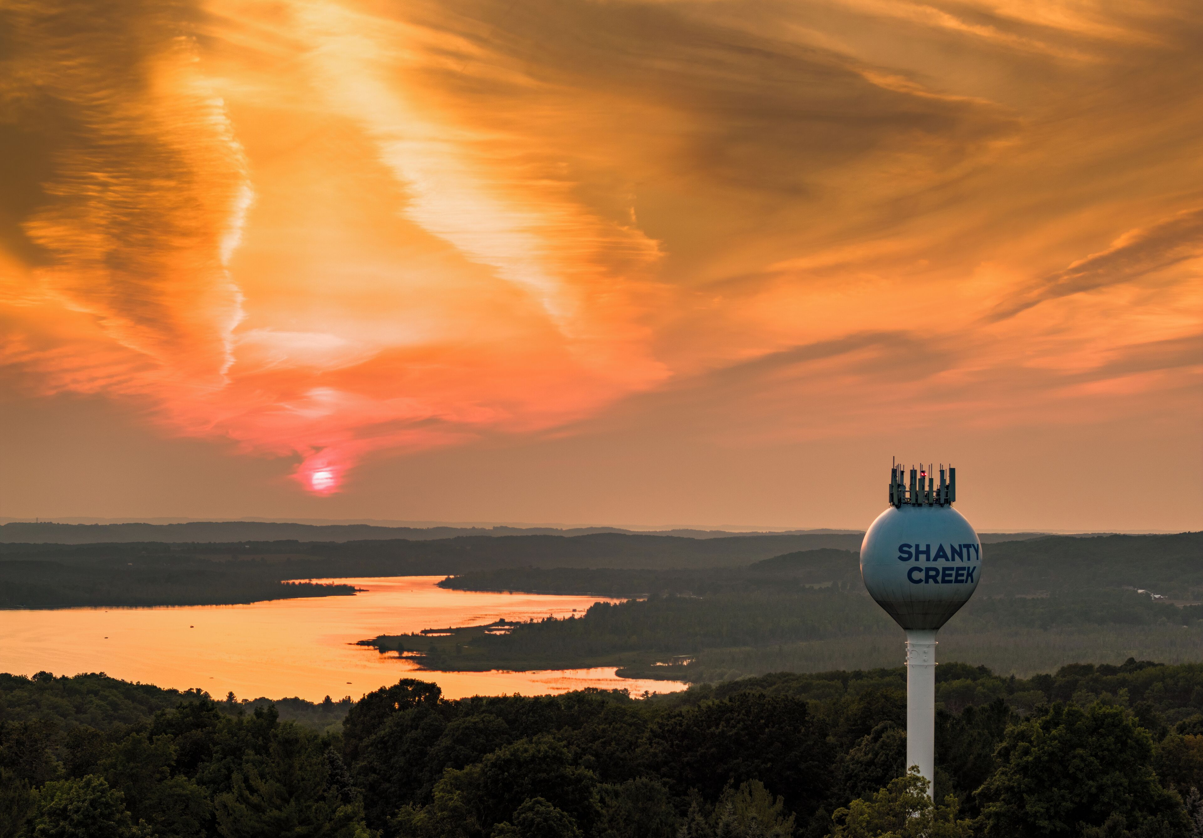 A water tower with "Shanty Creek" written on it, surrounded by a forested landscape and a sunset over a body of water.