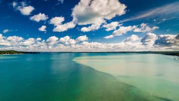 A wide, clear turquoise sea with a sandy beach and a distant tree line under a partly cloudy sky.