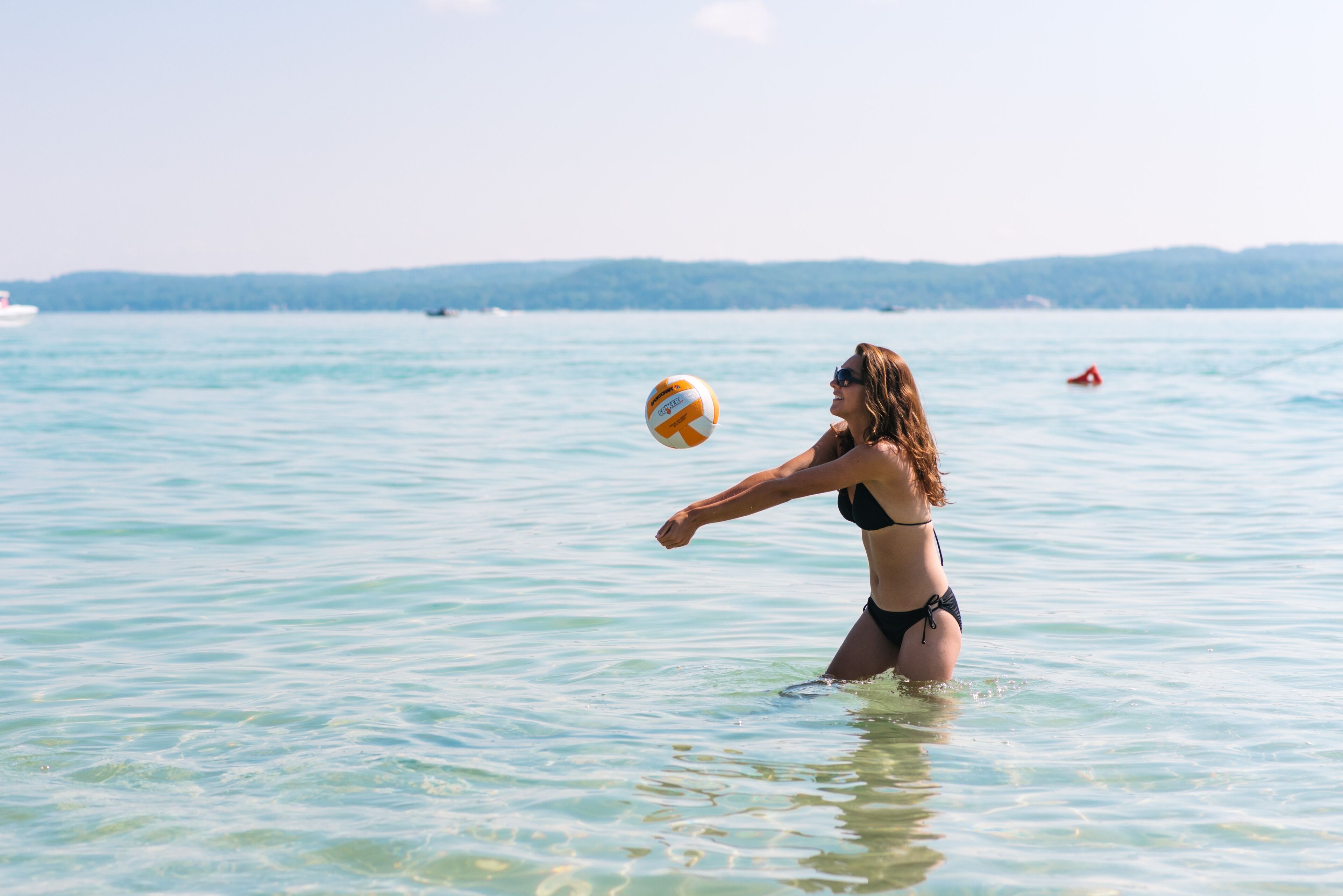A person playing volleyball in a body of water with a distant shoreline and boats.