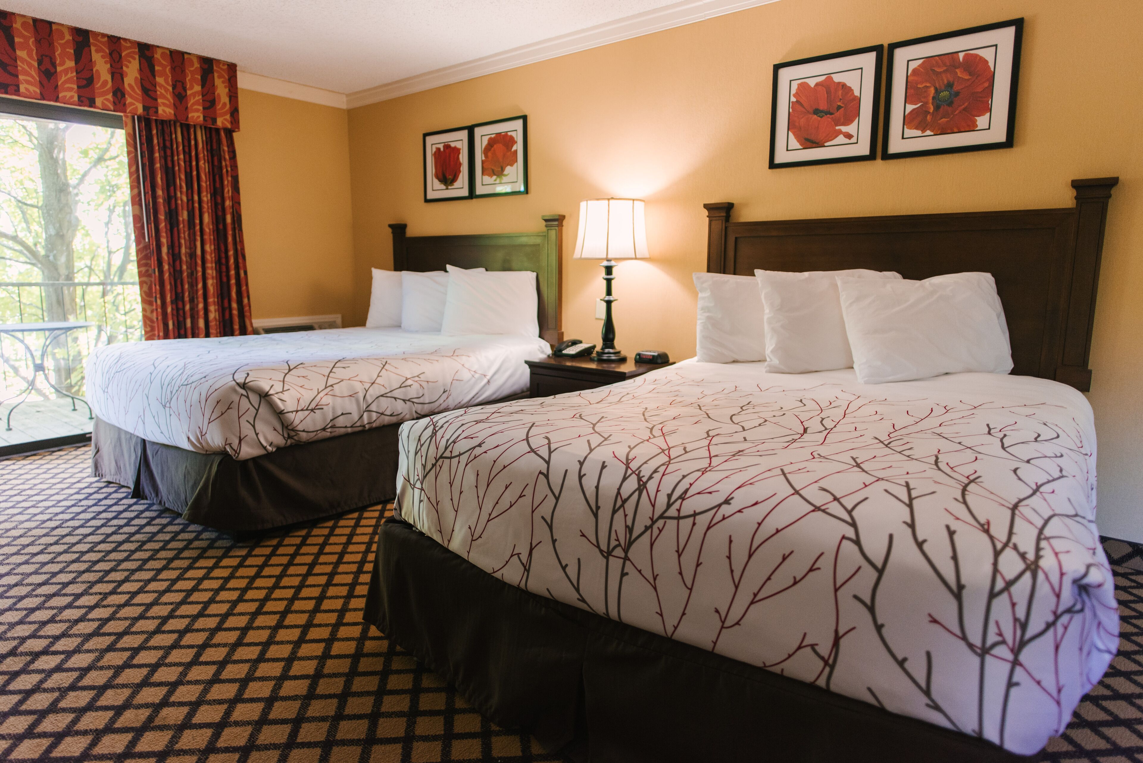 Une chambre d’hôtel avec deux lits, une fenêtre avec des rideaux, une table de chevet avec une lampe et des œuvres d’art florales encadrées accrochées au mur.