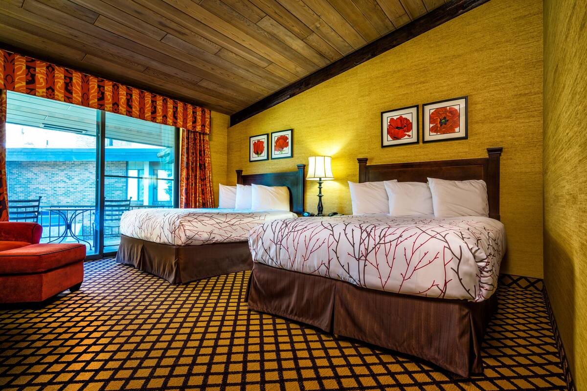 Habitación de hotel con dos camas, un sillón rojo, piso estampado y un balcón con vistas.