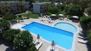 Una piscina con sillas de descanso y sombrillas, rodeada de vegetación y edificios.