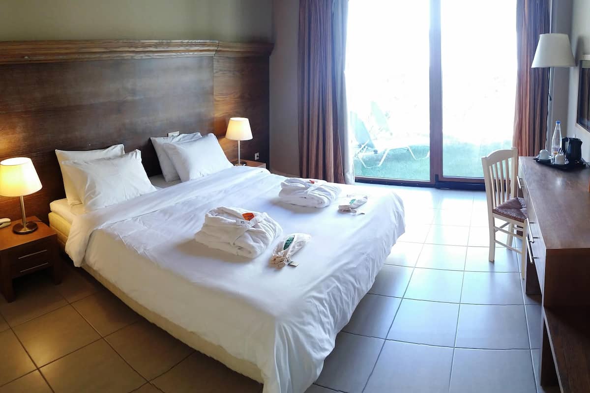 A hotel room with a large bed, two bedside lamps, a chair, a desk with a lamp, and a view of the outdoors through a large window.