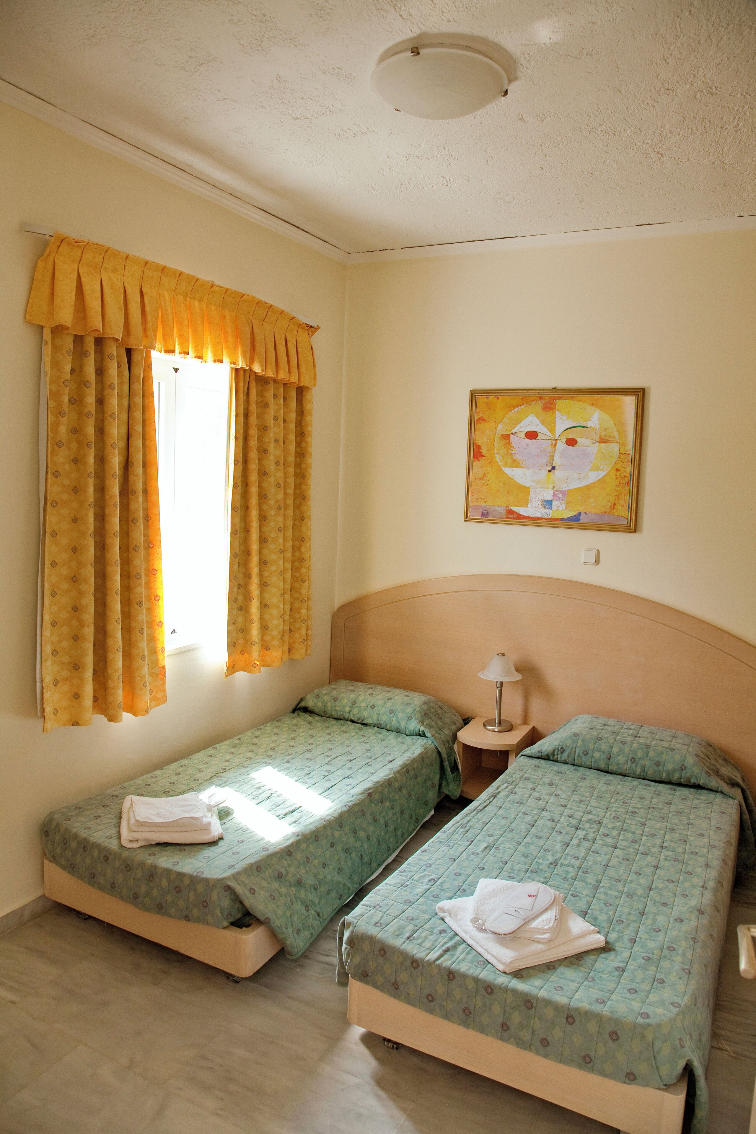 Una habitación con dos camas individuales, una mesita de noche con lámpara, una ventana con cortinas amarillas y un cuadro en la pared.