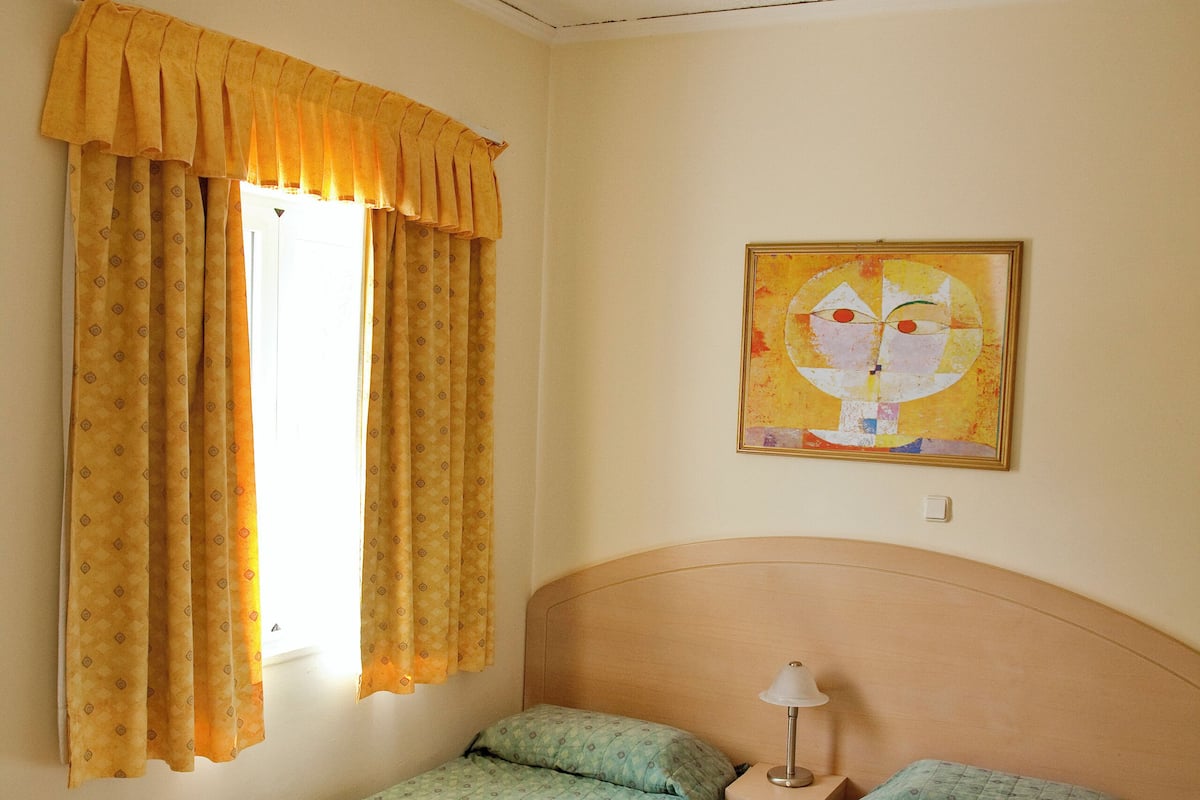 Una habitación con dos camas individuales, una mesita de noche con lámpara, una ventana con cortinas amarillas y un cuadro en la pared.