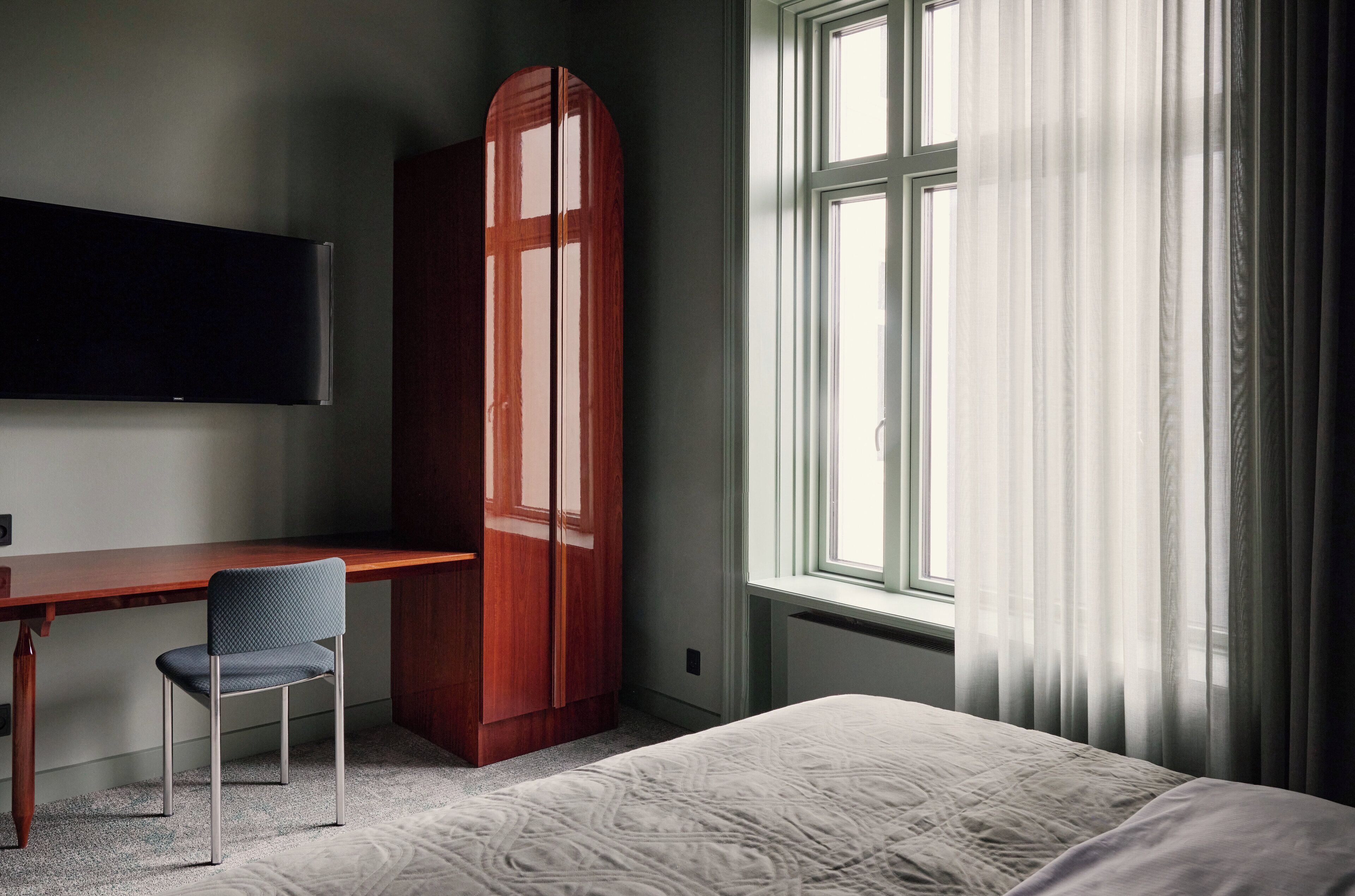 A bedroom with a bed, a desk, a chair, a wardrobe, and a window with curtains.