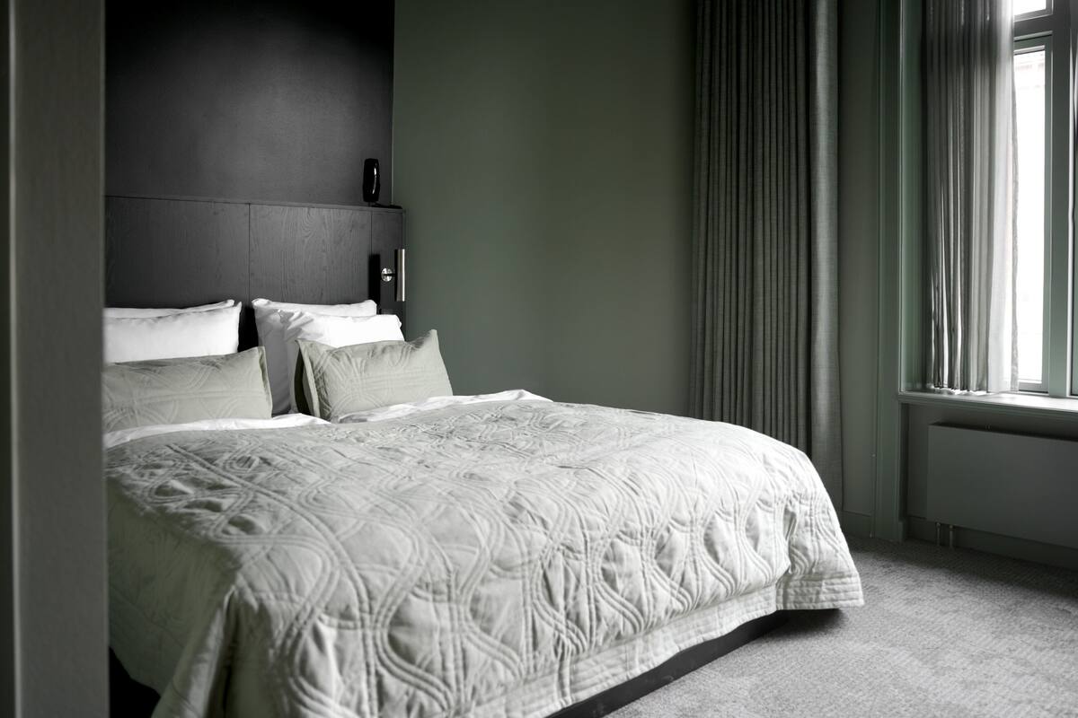 A modern hotel room with a large bed, a dark headboard, and a view of the outdoors through a window with curtains.