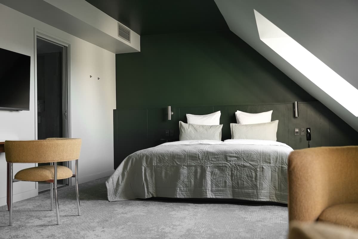 A modern bedroom with a sloped ceiling, a bed with white bedding, a chair, and a TV mounted on the wall.