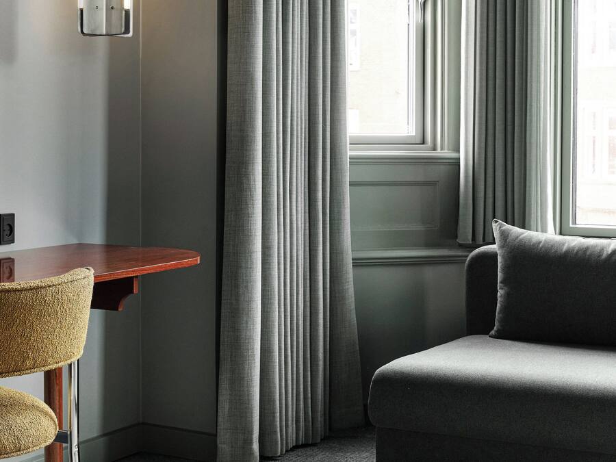 A modern hotel room with a wooden desk, a cushioned armchair, a sofa, and a large window with curtains.