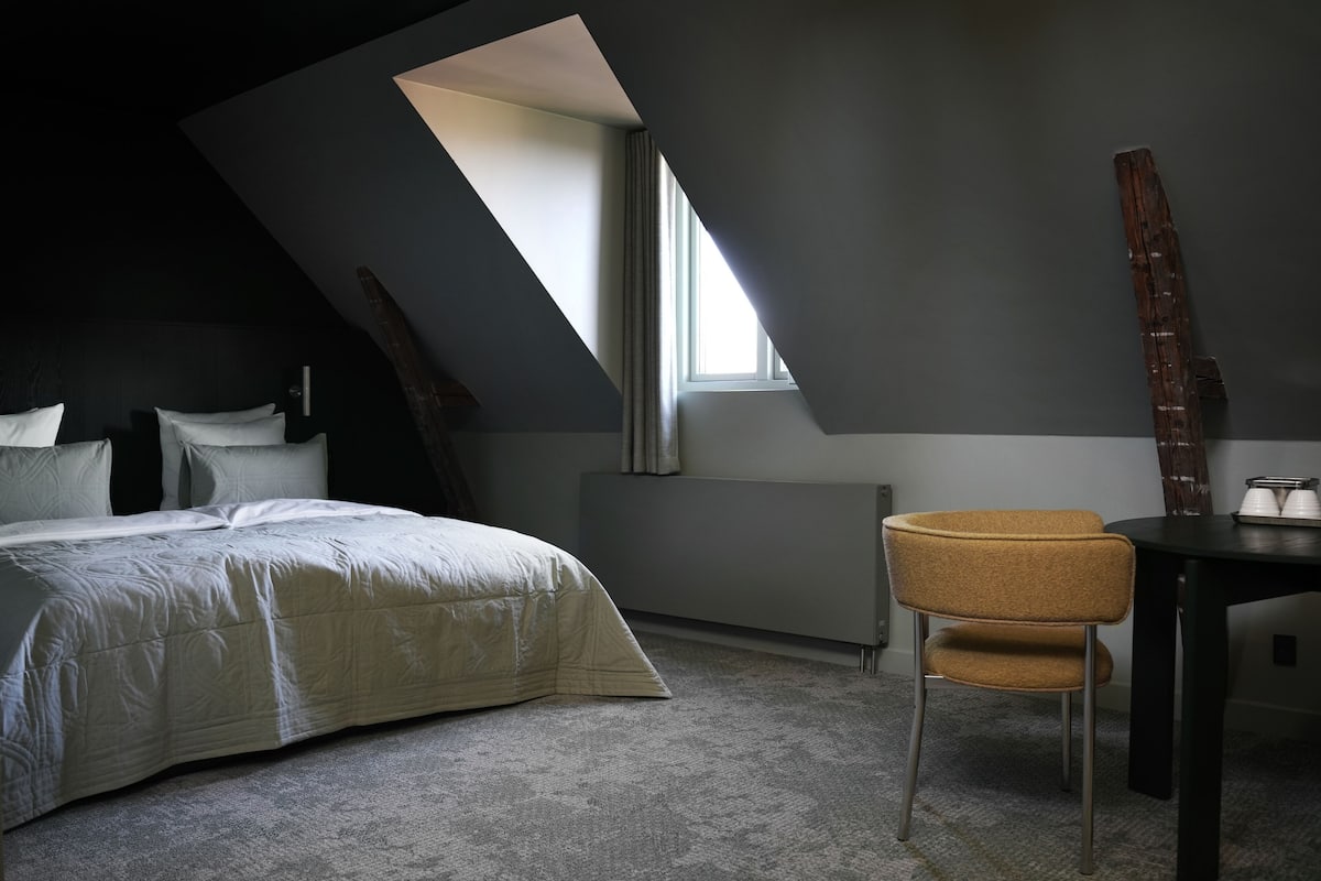 A bedroom with a bed, a chair, a table, a window, and a radiator.