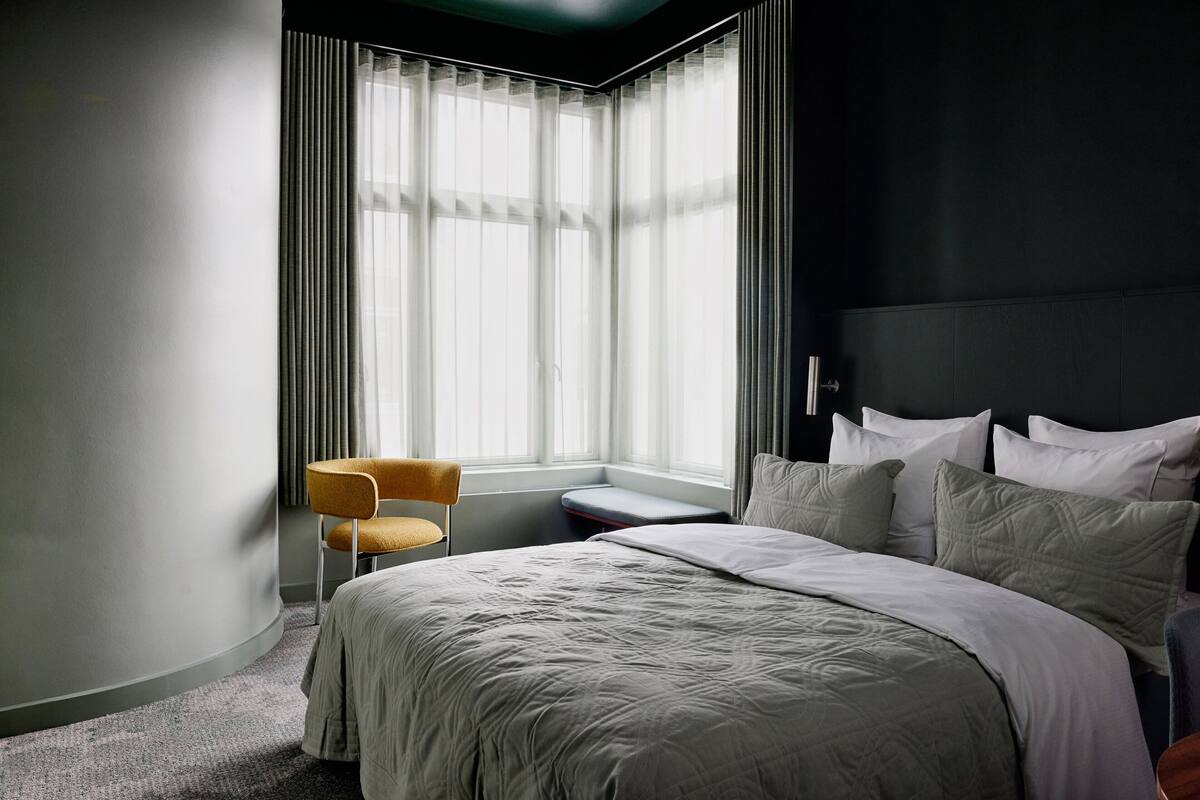 A modern hotel room with a large bed, a small seating area, and a window with sheer curtains.