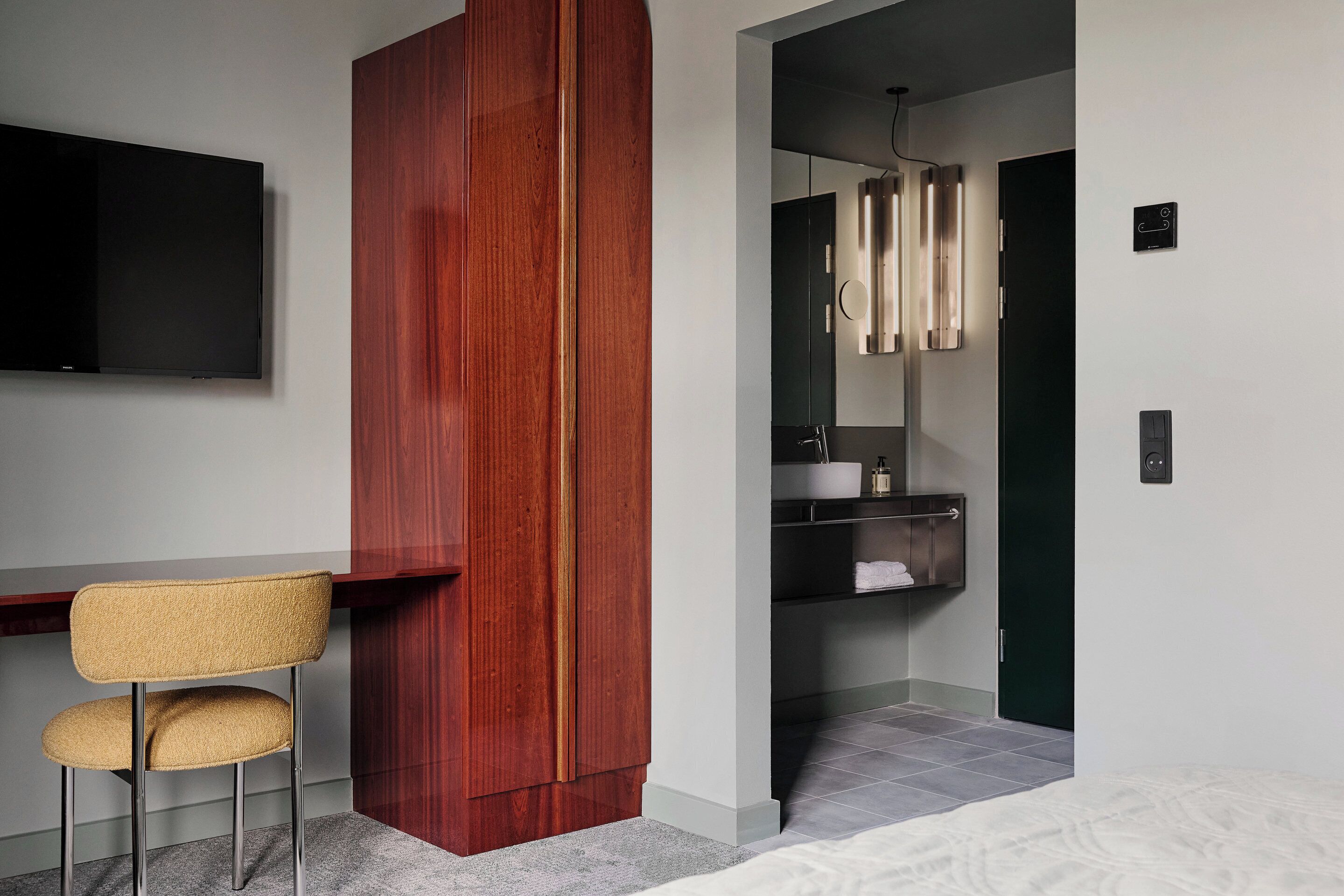 A modern room with a wooden wardrobe, a desk with a chair, a television, a bathroom with a mirror, and a bed.