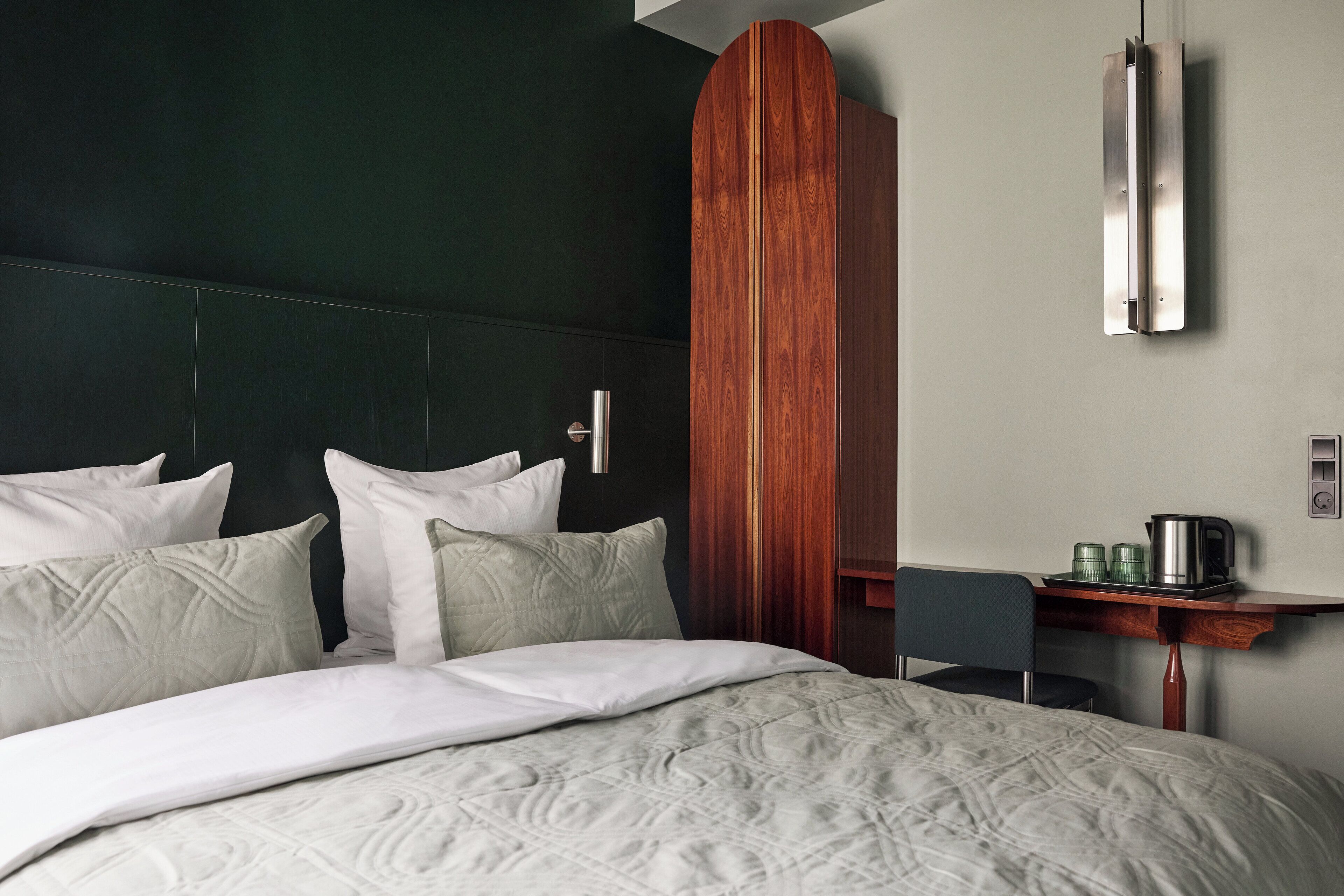 A hotel room with a bed, pillows, a wooden wardrobe, a desk with a kettle, and a mirror.