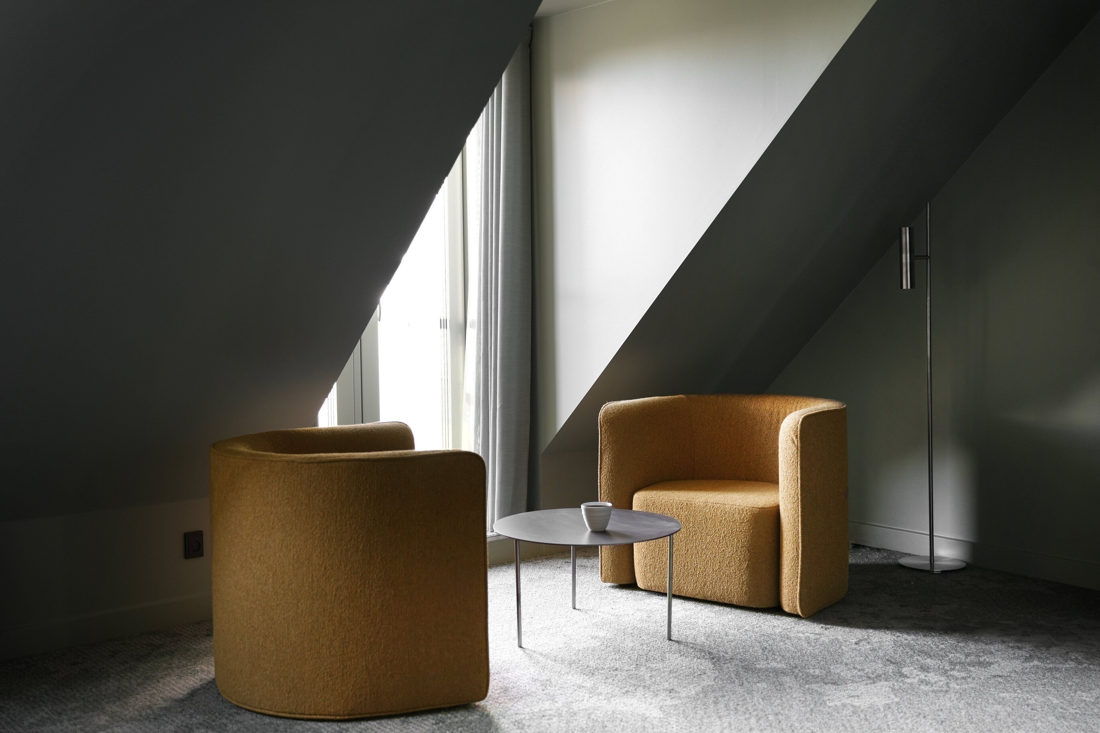 A modern loft bedroom with a large window, a round coffee table, and two upholstered chairs.