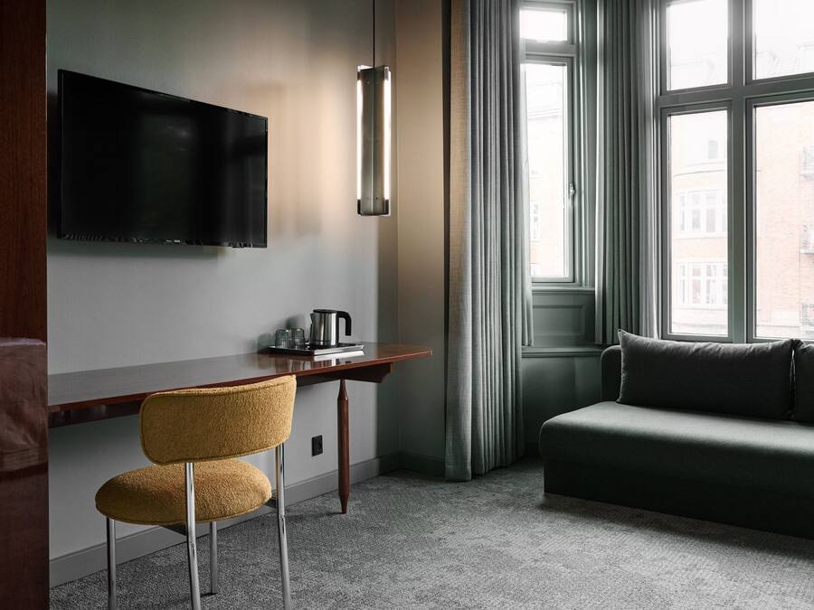 A modern hotel room with a flat-screen TV mounted on the wall, a wooden desk with a kettle, a yellow chair, a grey sofa, and a large window with curtains.
