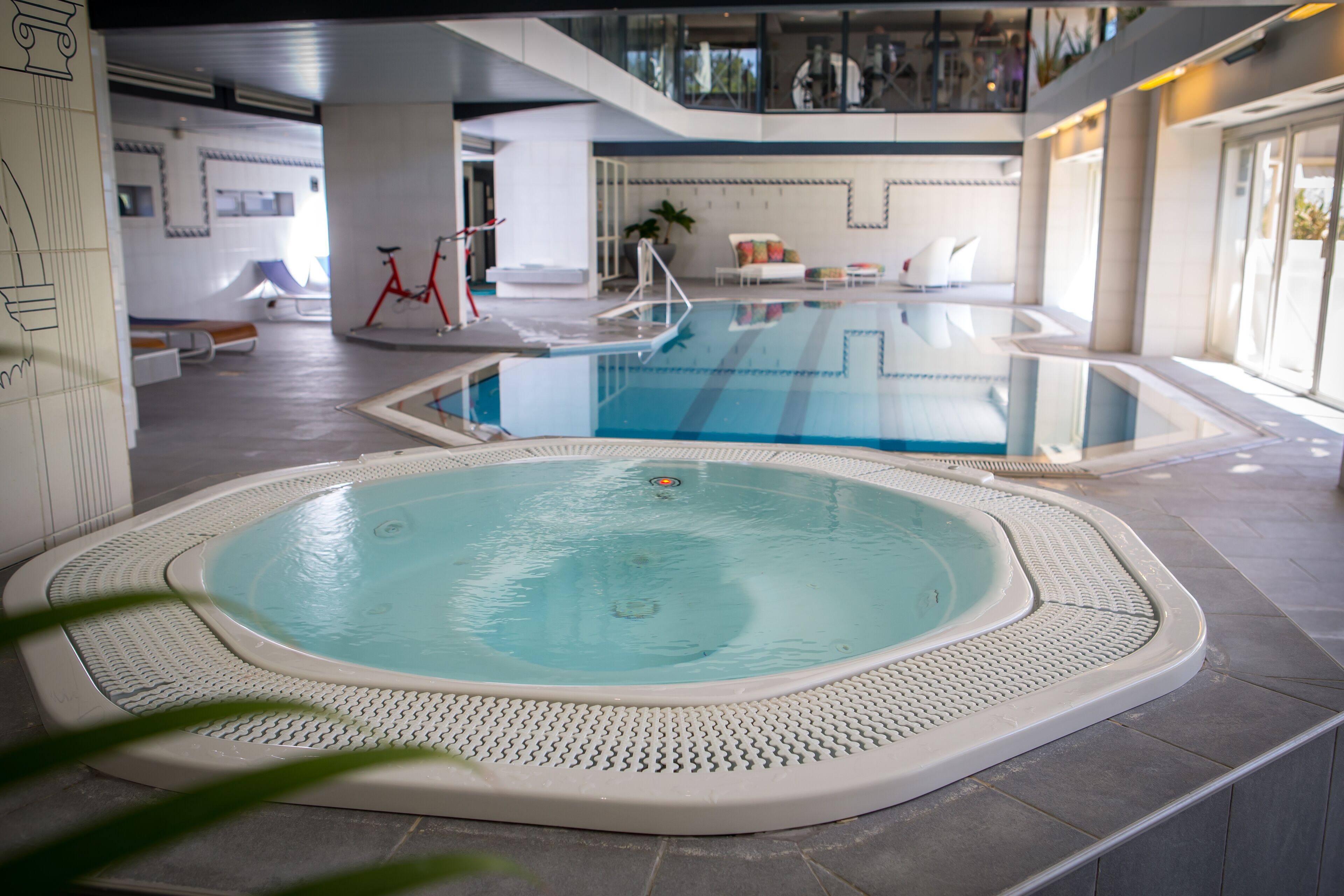 An indoor swimming pool with a hot tub, surrounded by a tiled floor and a white, textured border.