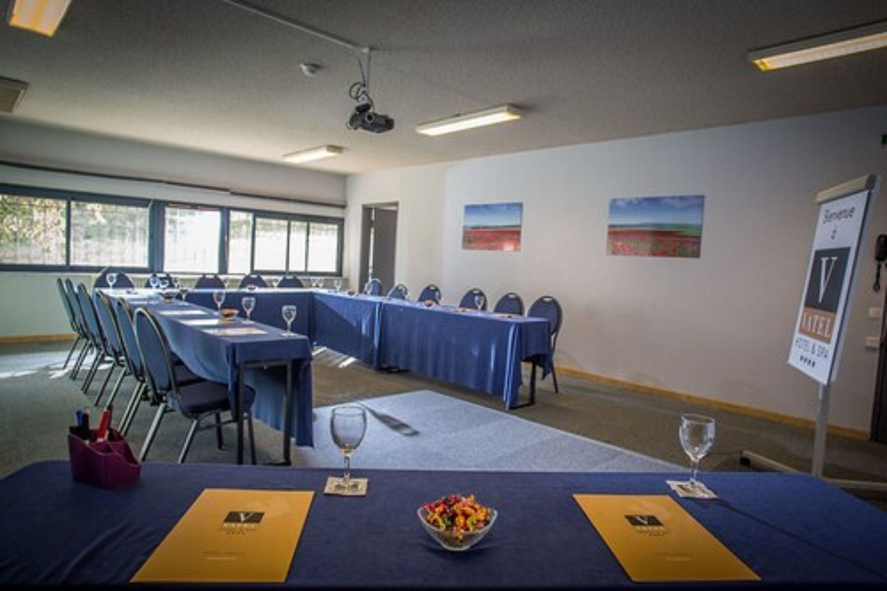 A conference room with long tables, chairs, and a projector. There are paintings on the walls and a sign indicating a hotel room.