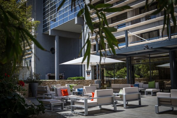 An outdoor seating area with white sofas and tables, a sign for 'Vatel', and a modern building in the background.