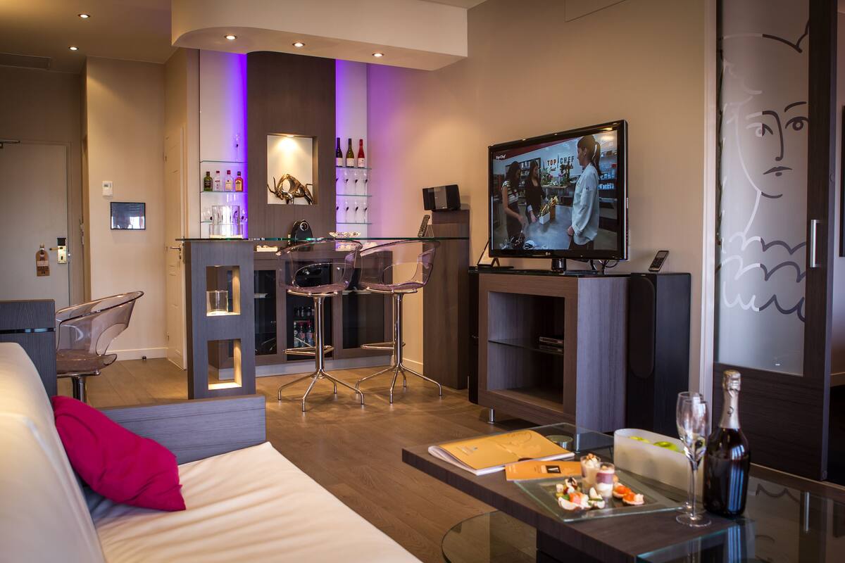 Una habitación de hotel moderna con sofá, una mesa de comedor de vidrio con comida, un televisor, una zona de bar con botellero y una chimenea.