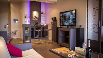 A modern hotel room with a sofa, a glass dining table with food, a TV, a bar area with a wine rack, and a fireplace.