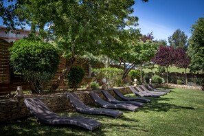 A row of sun loungers aligned along a stone wall in a well-maintained garden area with various trees and shrubs.