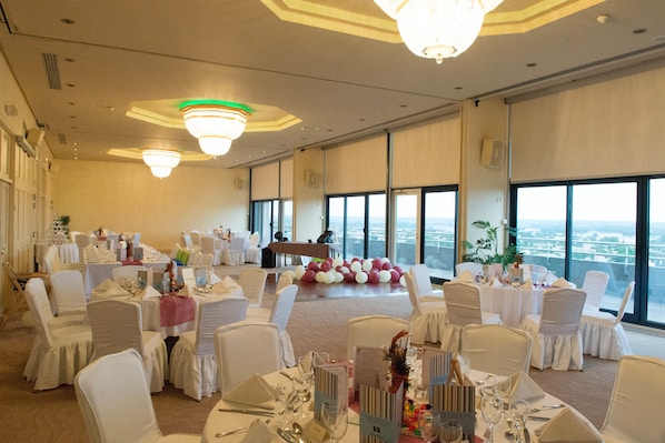 A banquet hall with round tables set for dining, large windows offering a city view, and a stage with a podium.