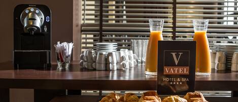 Una mesa con croissants, tazas de café y un dispensador de jugo.