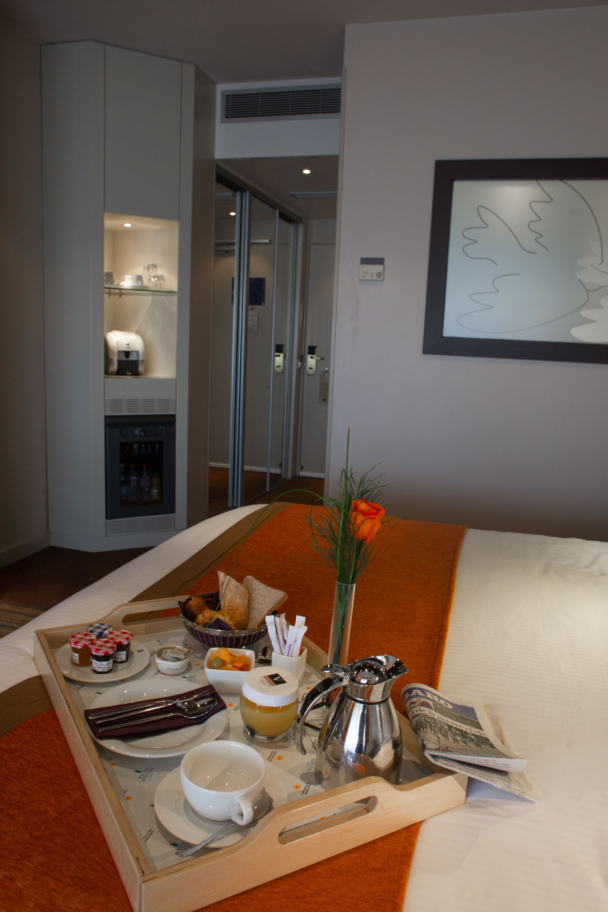Habitación de hotel con bandeja de desayuno, cafetera, un florero con flores y una obra de arte enmarcada en la pared.