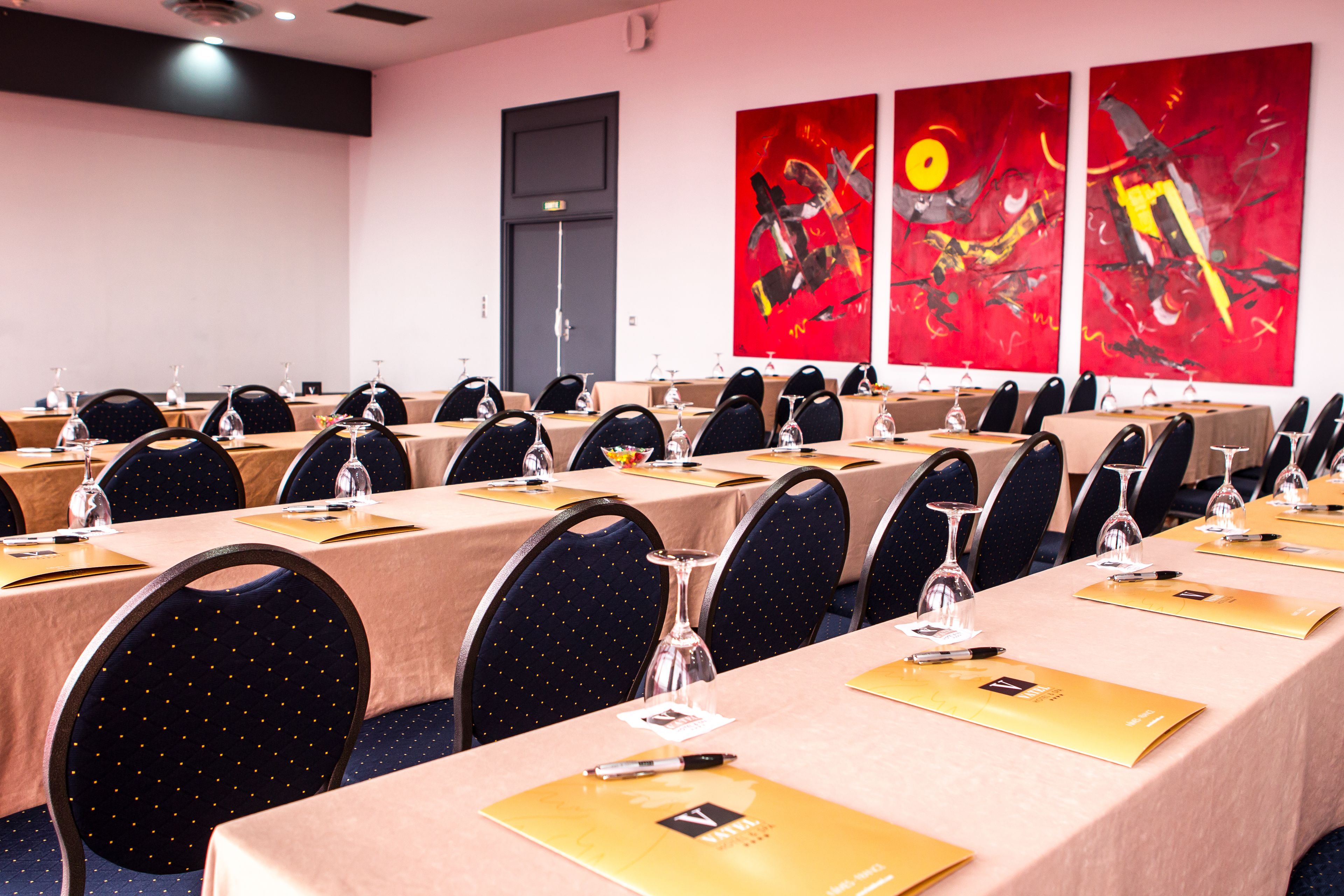 A conference room with multiple tables, each with a name card holder, a pen, and a glass. The room has a modern design with a dark ceiling and a door in the background.