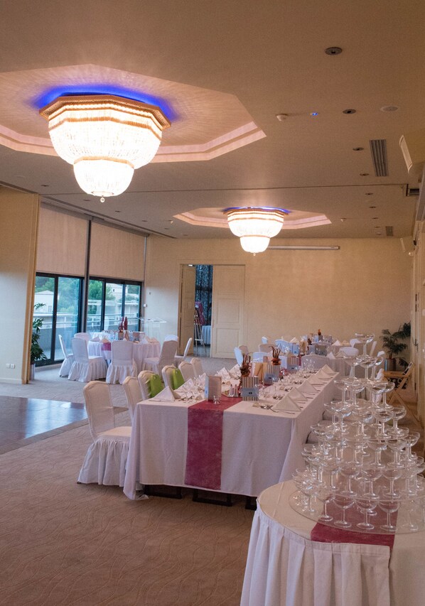 A banquet hall with tables set for a formal event, featuring a chandelier and large windows.