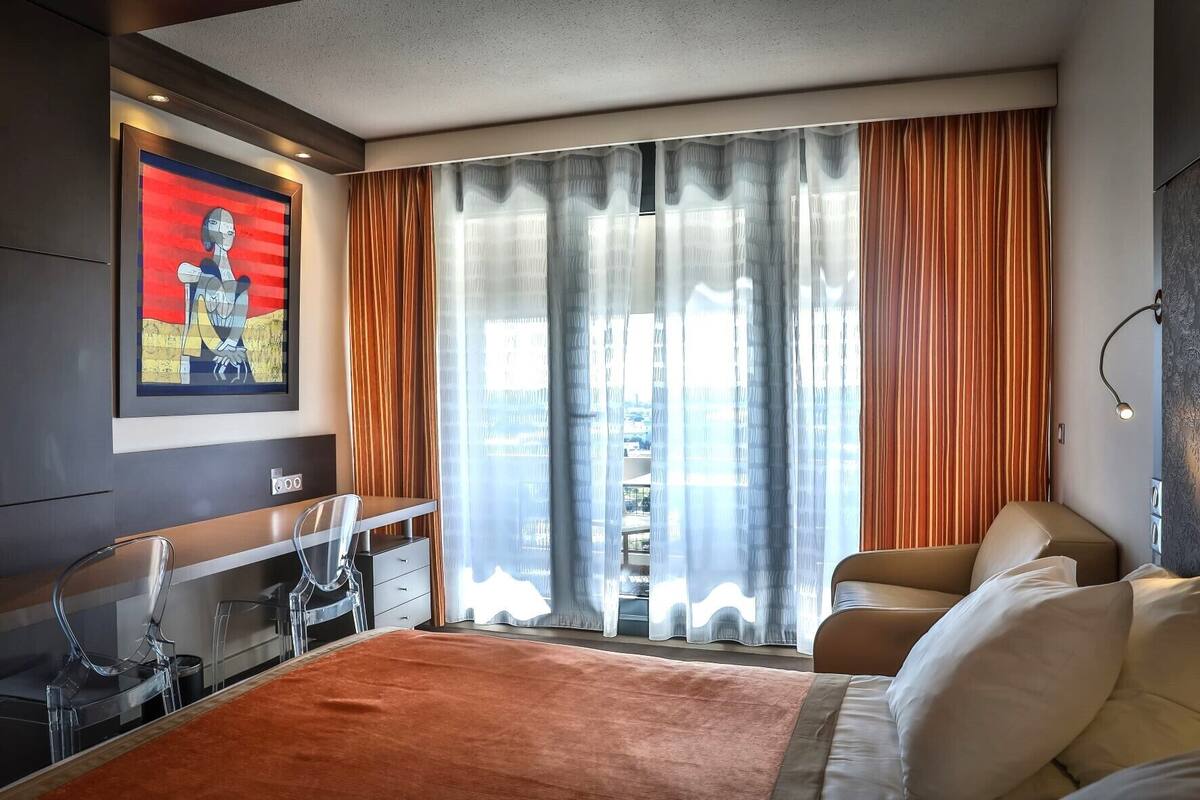 Un hotel moderno con una cama grande, un escritorio con silla, un cuadro en la pared y vistas a través de cortinas traslúcidas.