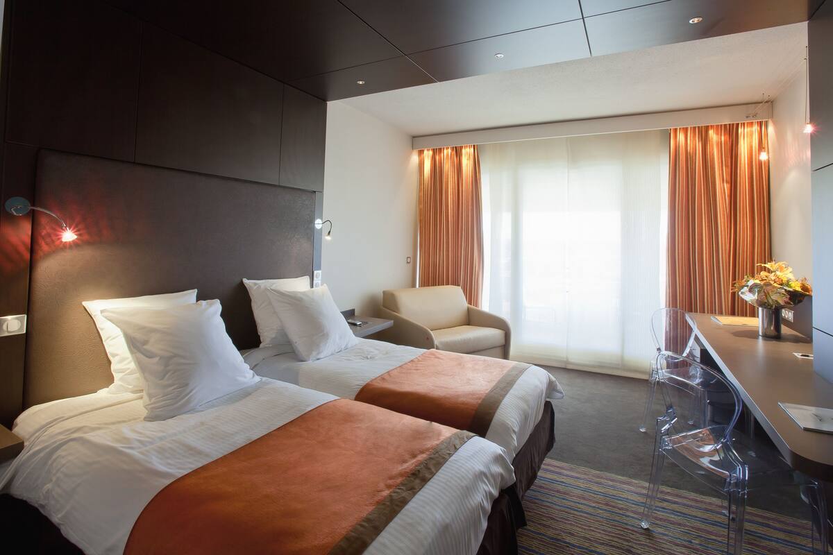 Habitación de hotel con dos camas, un escritorio, una silla y un ventanal con cortinas.