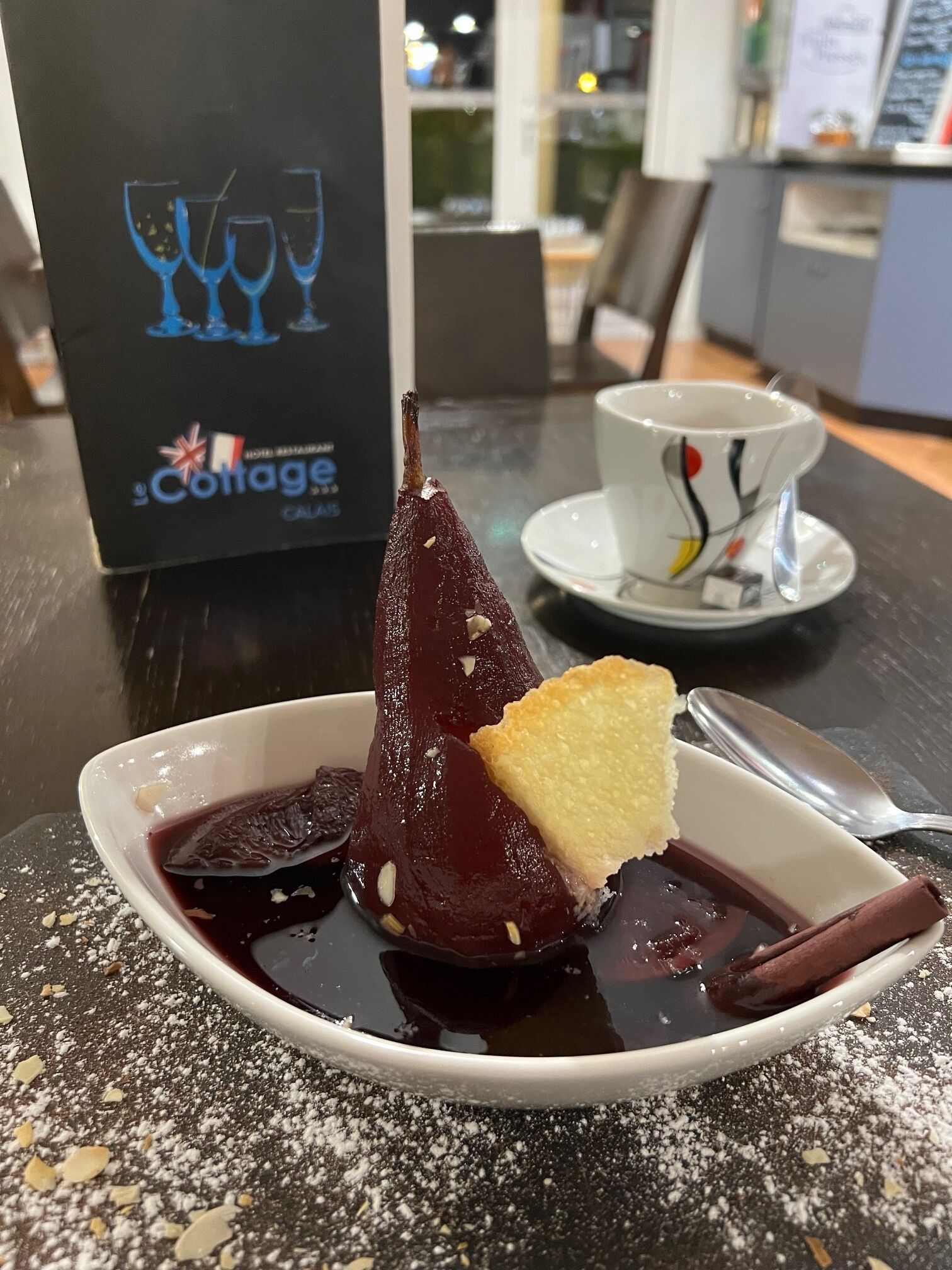 Un plato de postre con un dulce morado oscuro, una porción de pastel y una rama de canela sobre una mesa con motas. Al fondo, hay un atril con un logotipo y vasos.