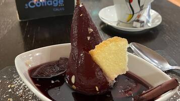 Un plato de postre con un dulce morado oscuro, una porción de pastel y una rama de canela sobre una mesa con motas. Al fondo, hay un atril con un logotipo y vasos.