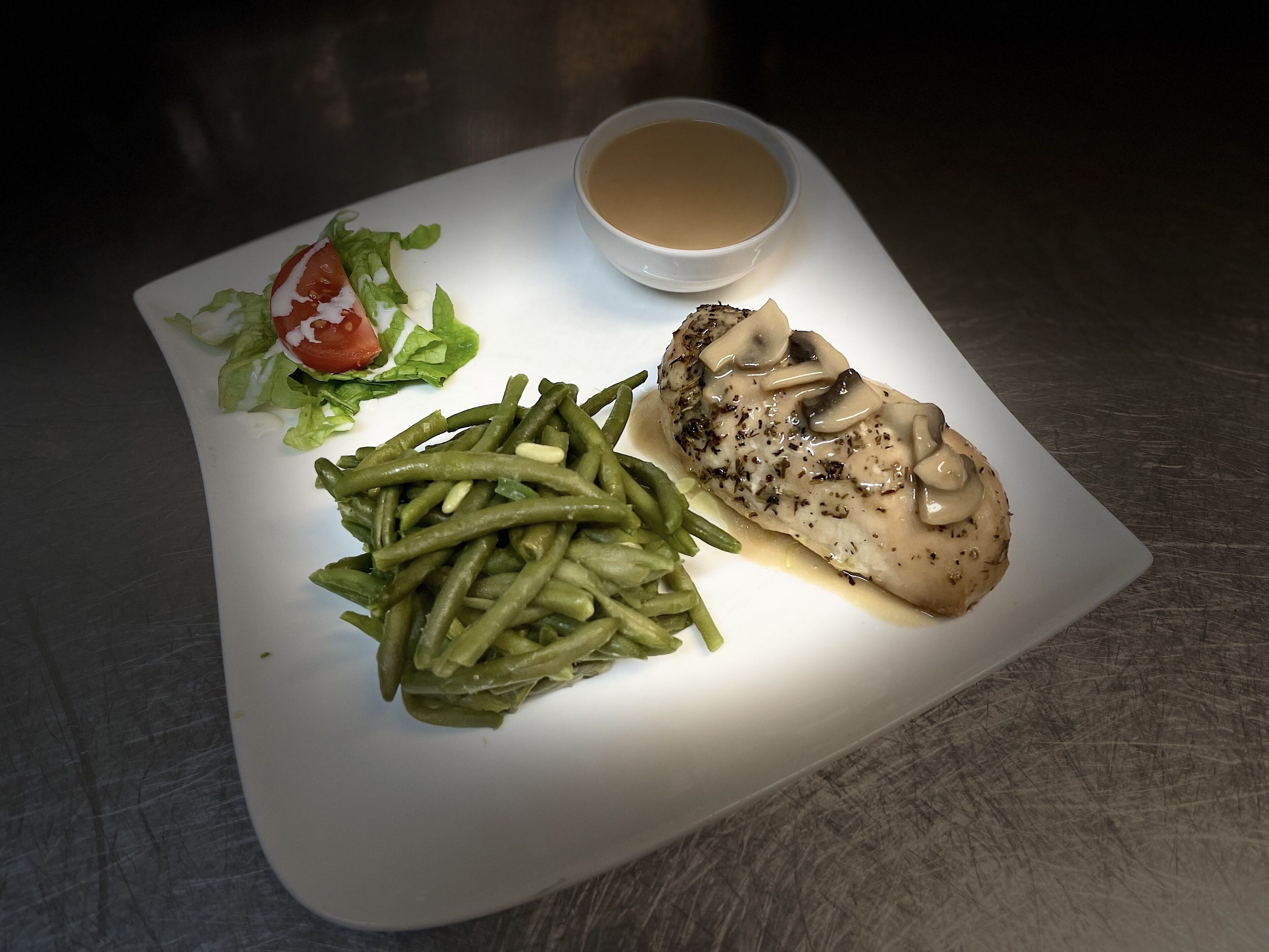 Un plato con un trozo de pollo cubierto de champiñones, judías verdes, una ensalada pequeña y una taza de sopa.