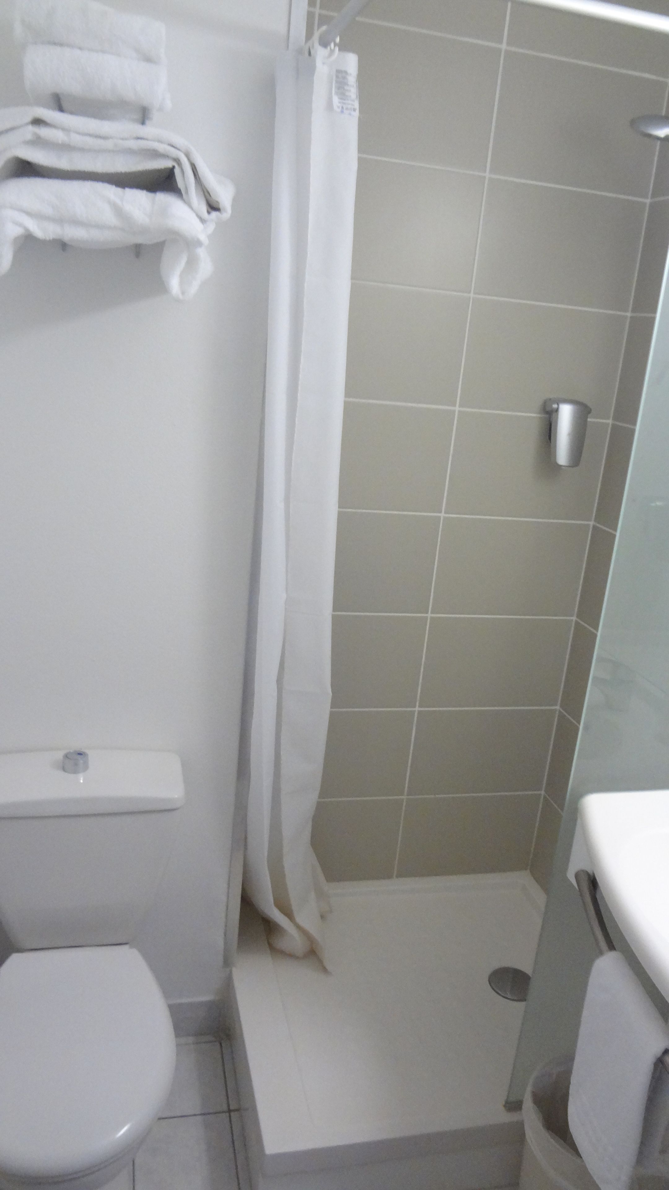 A small, enclosed shower area with a white curtain, a toilet, and a sink with a towel hanging on the wall.