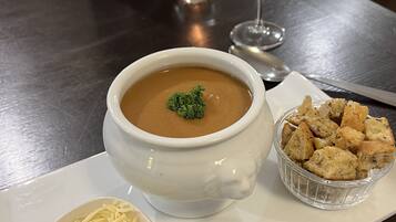 Una mesa de comedor con un tazón de sopa adornado con una ramita de perejil, un cuenco pequeño de queso rallado y una copa de vino blanco.