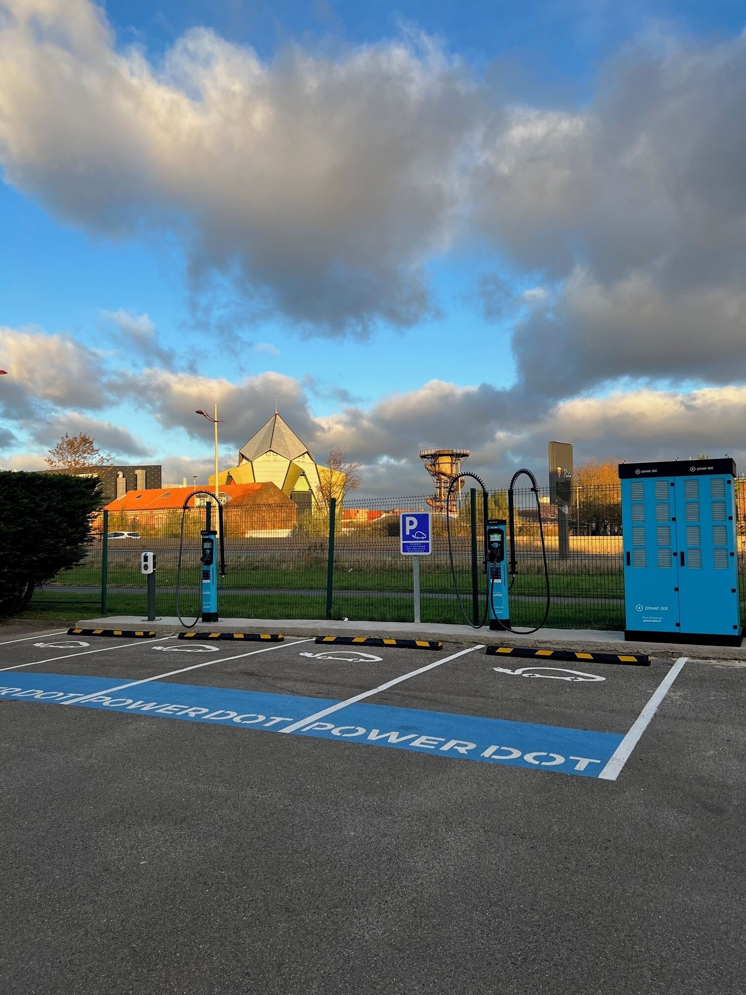 Ein Parkplatz mit Ladestationen für Elektrofahrzeuge, einem blauen Parkschild und einem eingezäunten Bereich mit einem Gebäude im Hintergrund.