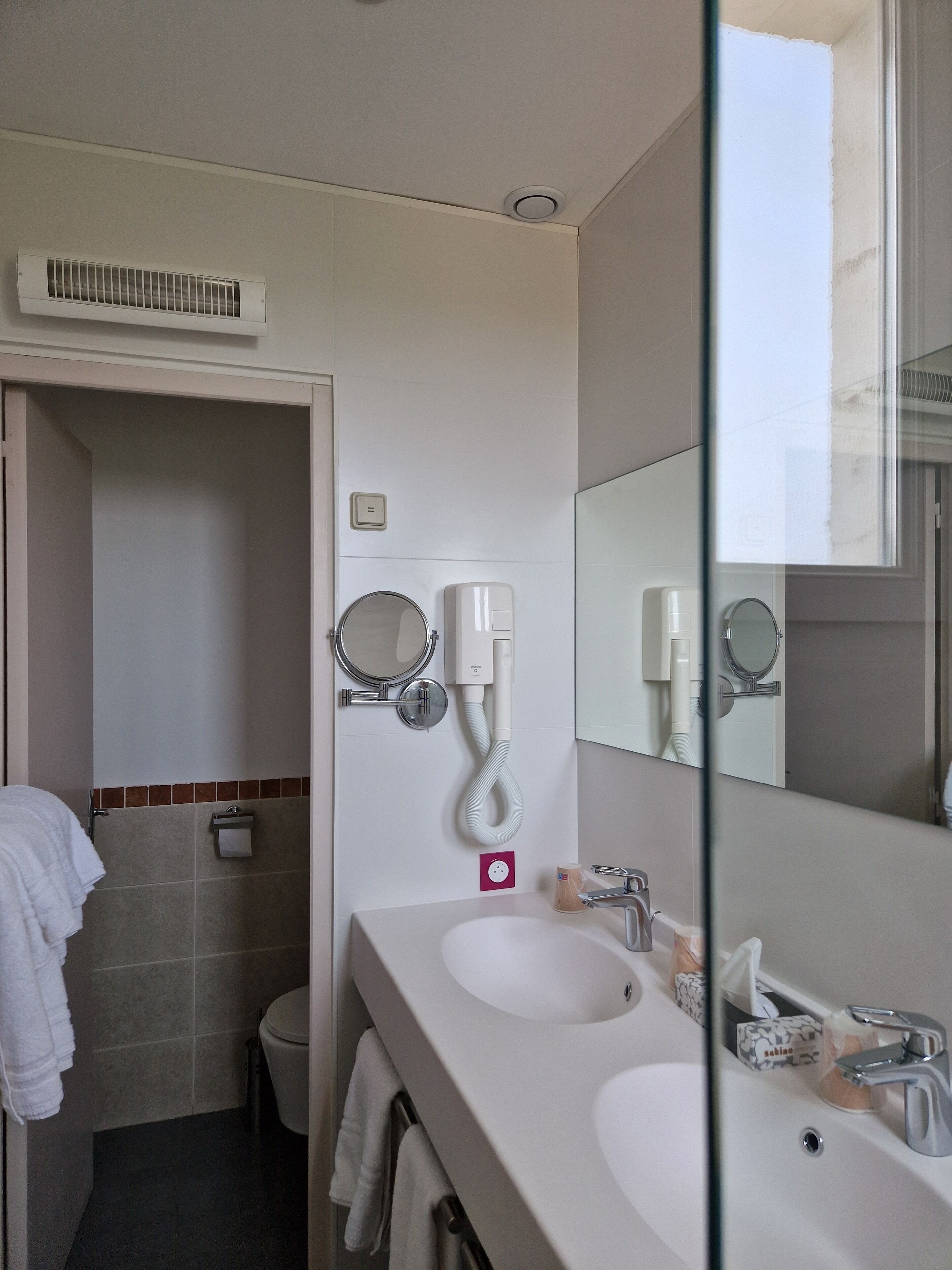 Une salle de bain moderne avec un lavabo blanc, un miroir et des toilettes. On y trouve un porte-serviettes avec des serviettes, un distributeur de savon et un porte-rouleau de papier toilette.