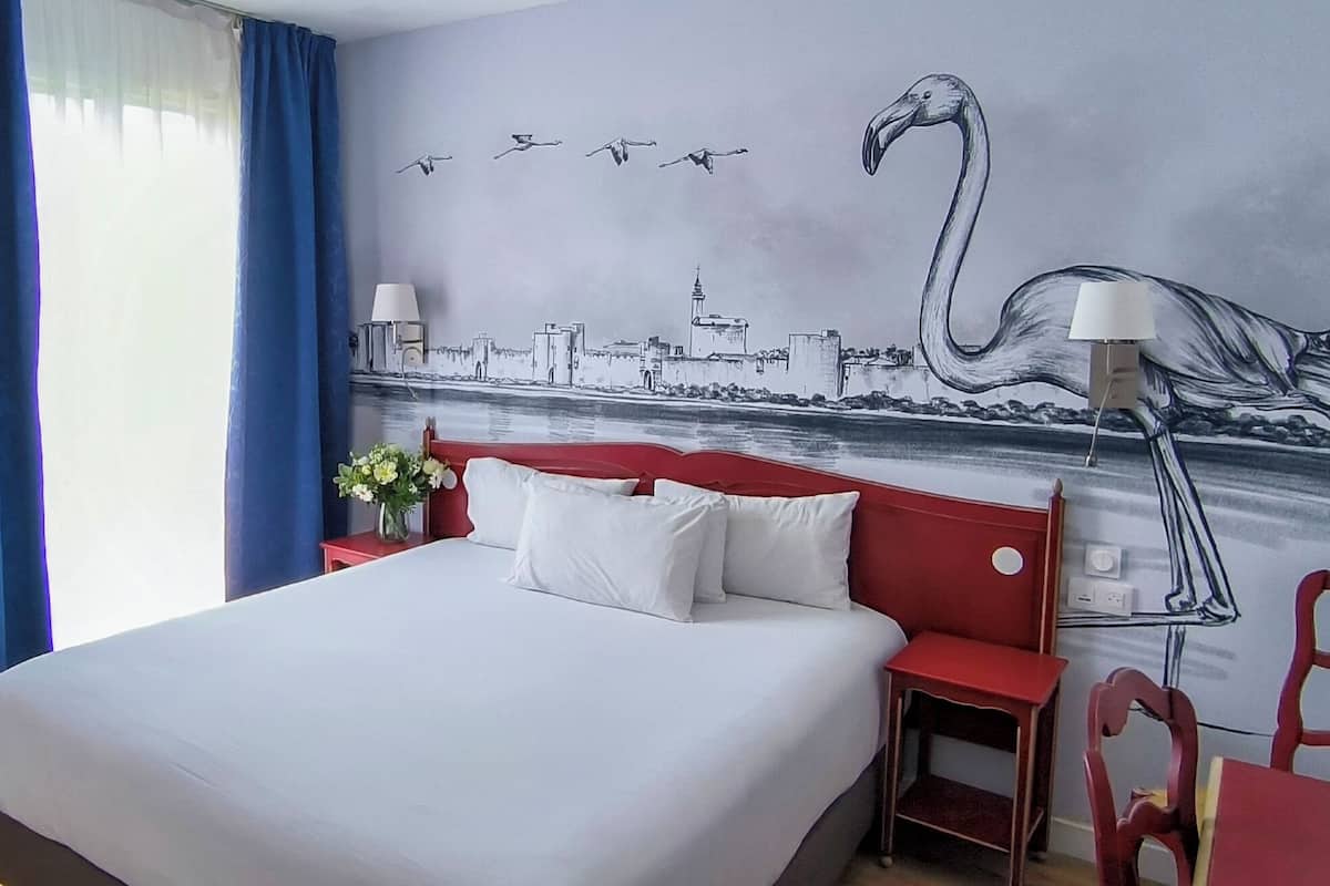 Una habitación de hotel con cama, televisión, cabecera roja, lámpara y un mural con un paisaje urbano y un flamenco.