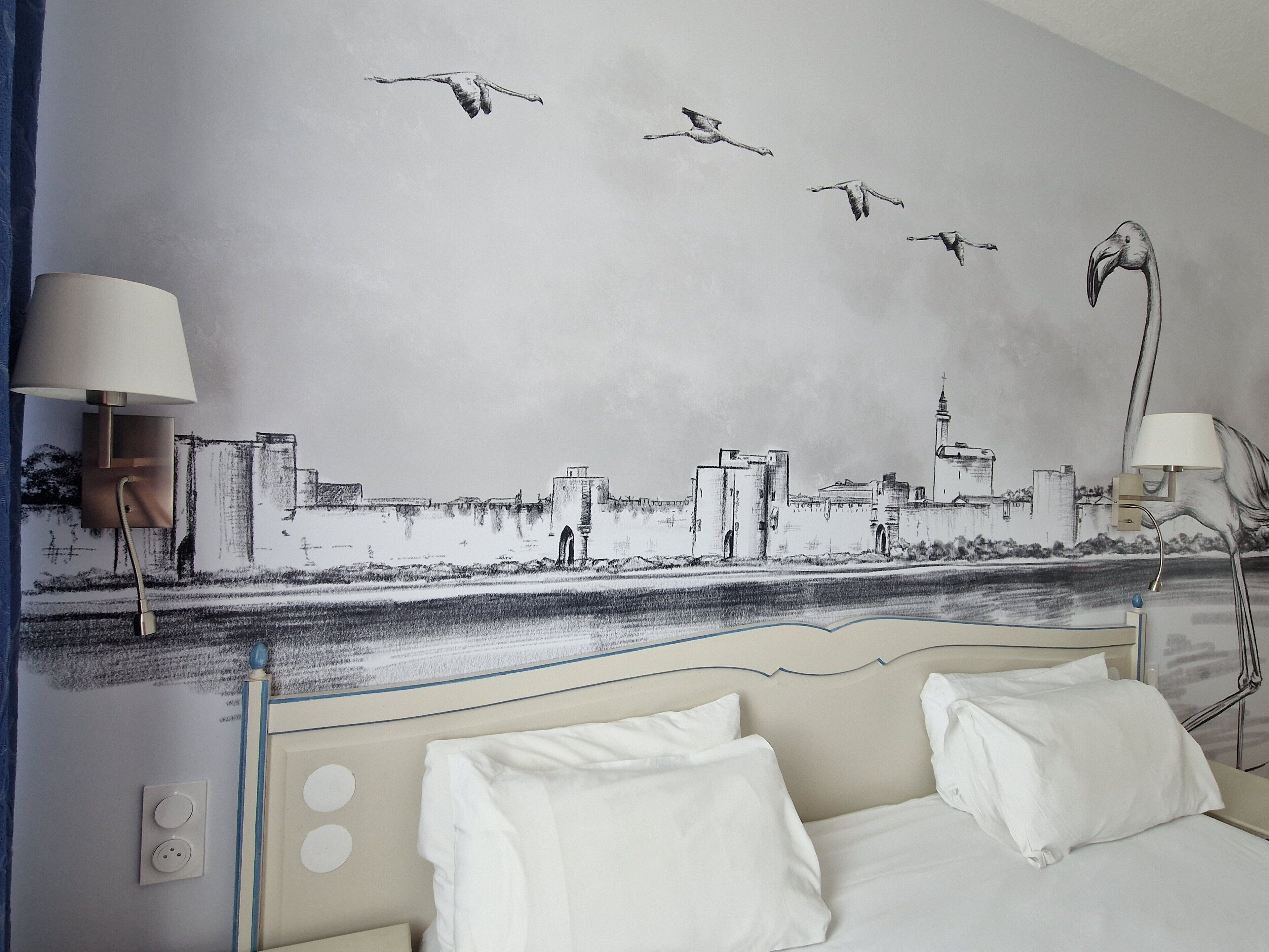 Habitación de hotel con una cama, dos lámparas de noche y un mural en la pared que muestra aves volando sobre un paisaje urbano.