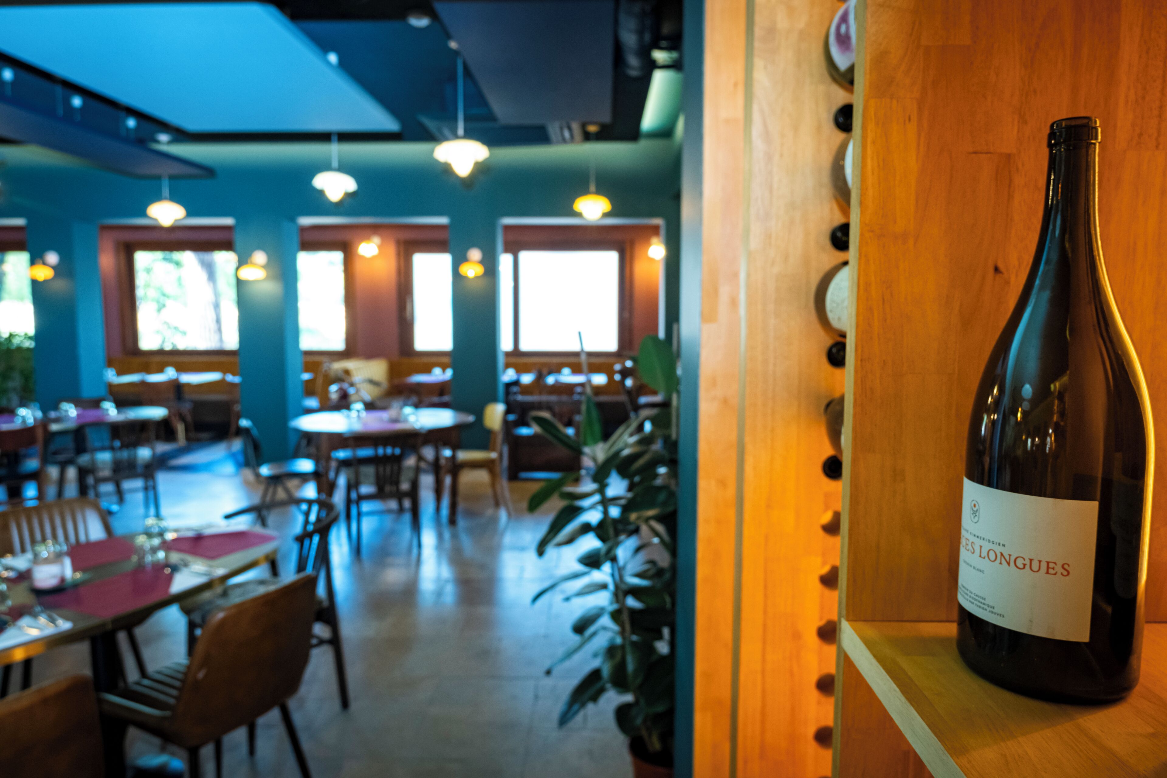 Interior de un restaurante con mesas, sillas y luces colgantes. Una botella de vino destaca en una estantería.