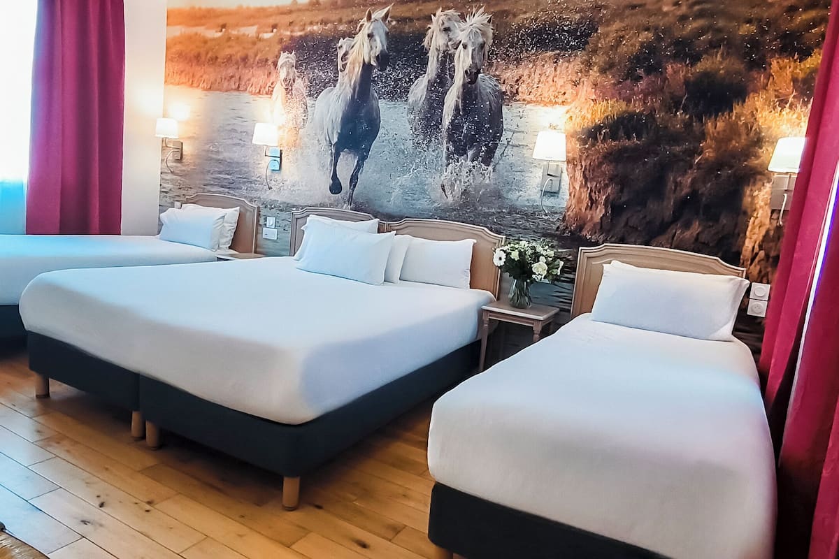 Habitación de hotel con tres camas, piso de madera, un mural en la pared y un florero con flores en la mesita de noche.