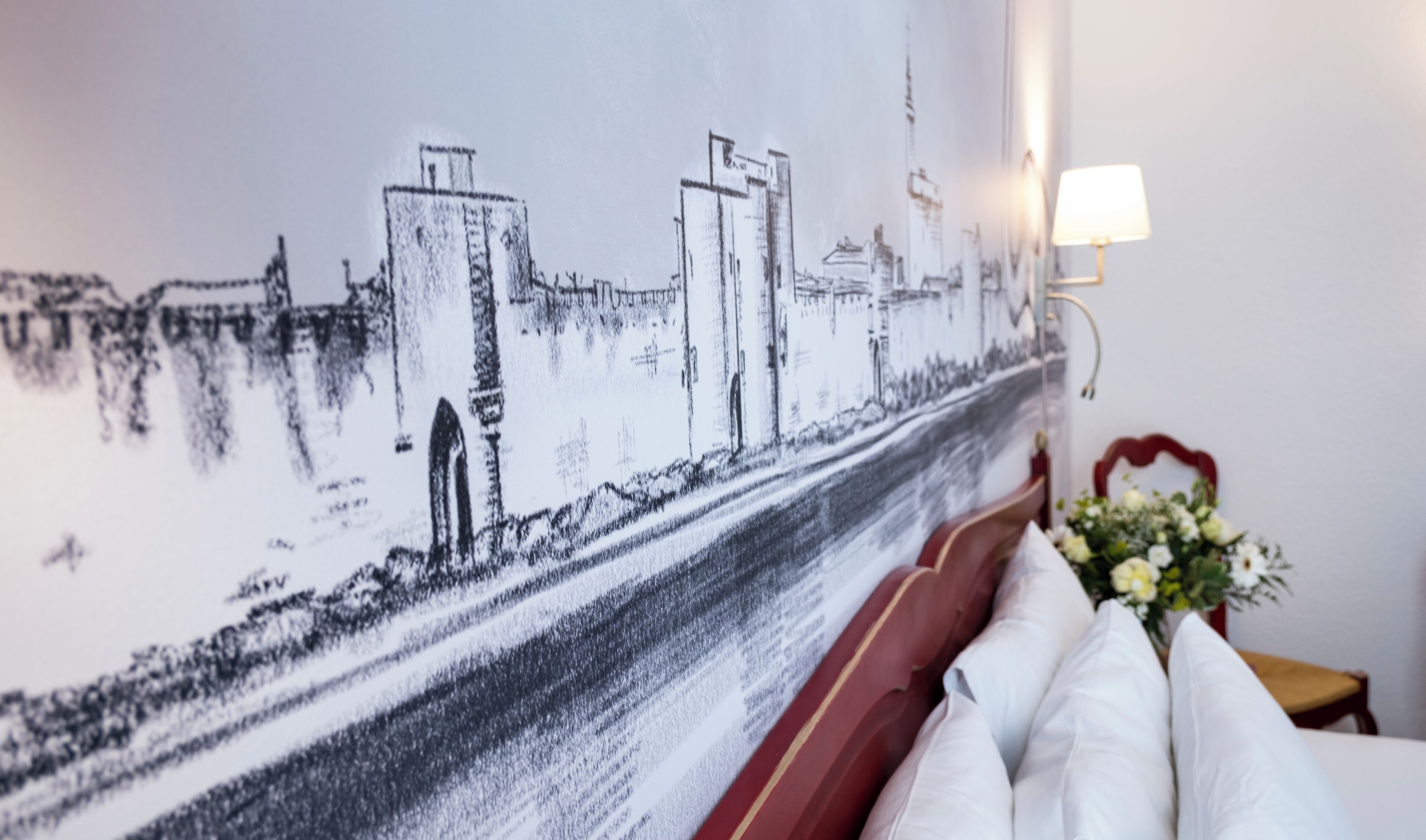 A wall mural of a cityscape, a bed with white pillows, and a bedside table with a lamp and flowers.